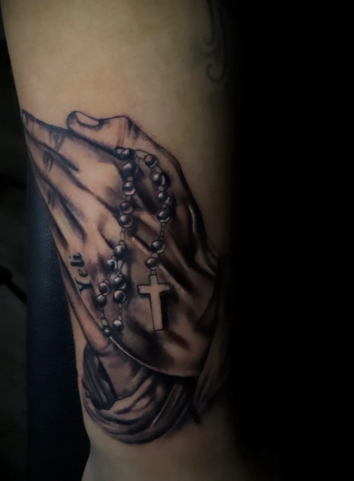 Black-and-grey Chicano-tradition praying hands clasping a rosary with cross on the upper arm.