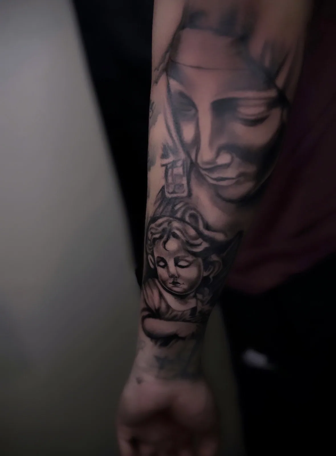 Black-and-grey realism forearm tattoo of the Virgin Mary with a cherub angel below.