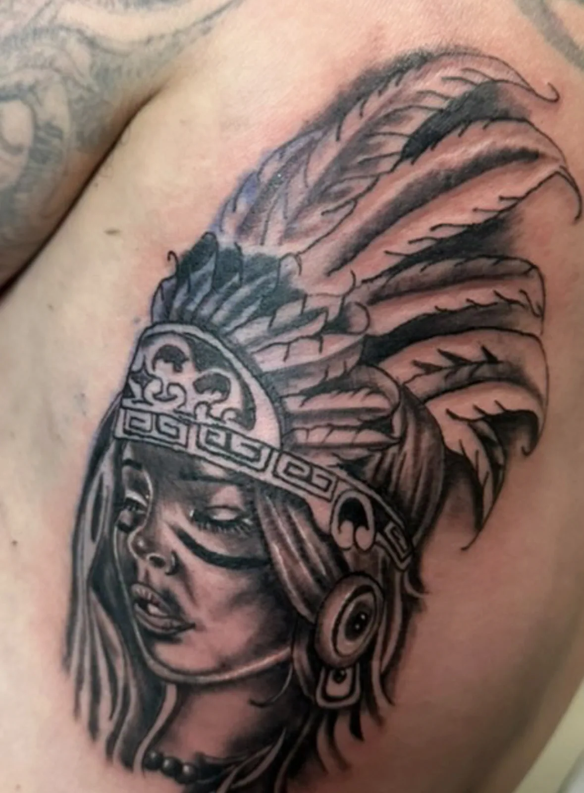 Black-and-grey Aztec warrior princess portrait with feathered headdress on the chest.