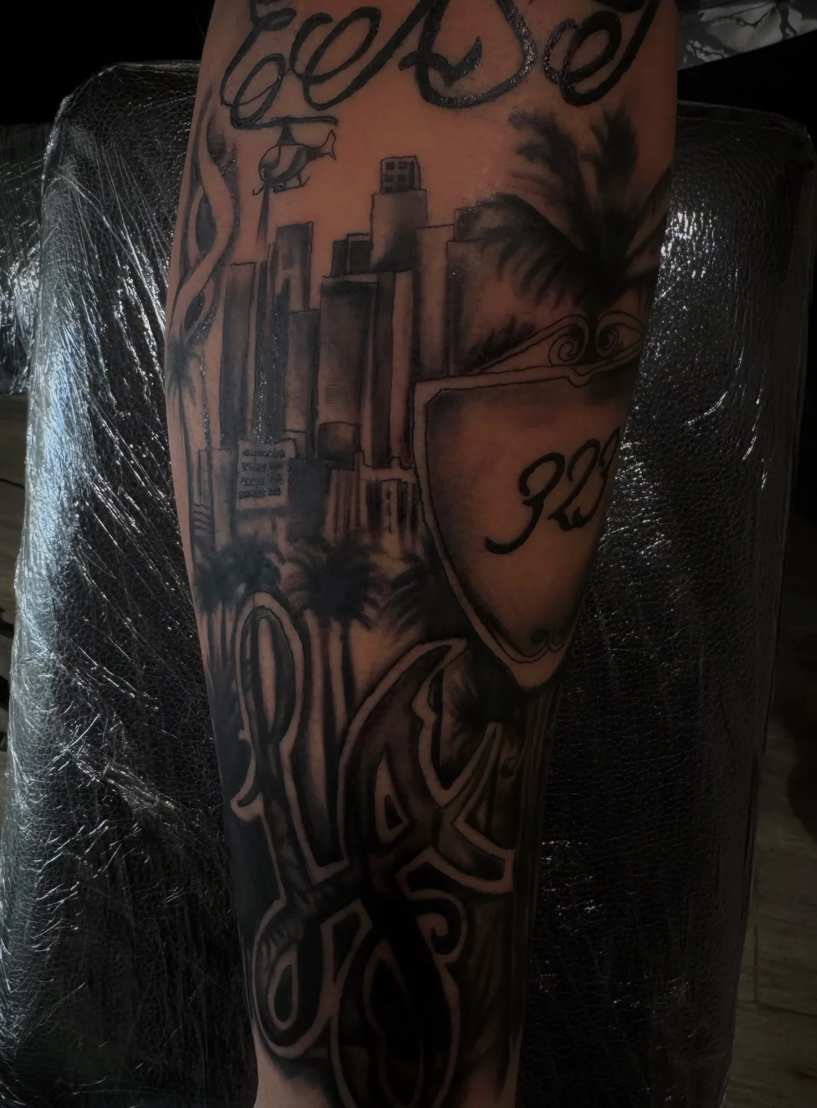 Black-and-grey Chicano-style forearm sleeve with LA skyline, palm trees, and script lettering.
